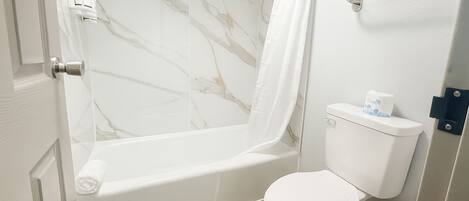 Baño con pared de ducha de mármol, una bañera blanca y un inodoro con dispensador de toallas de papel.
