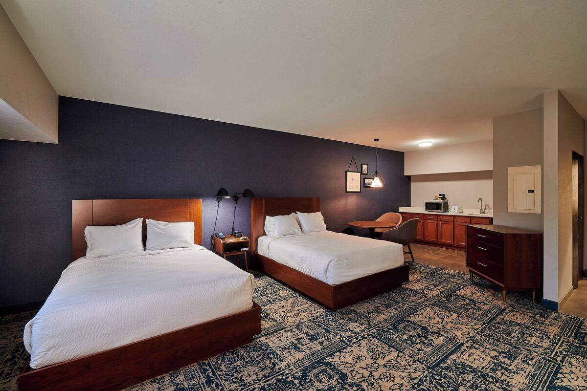 A hotel room with two beds, a desk, a chair, a microwave, and a small kitchenette.