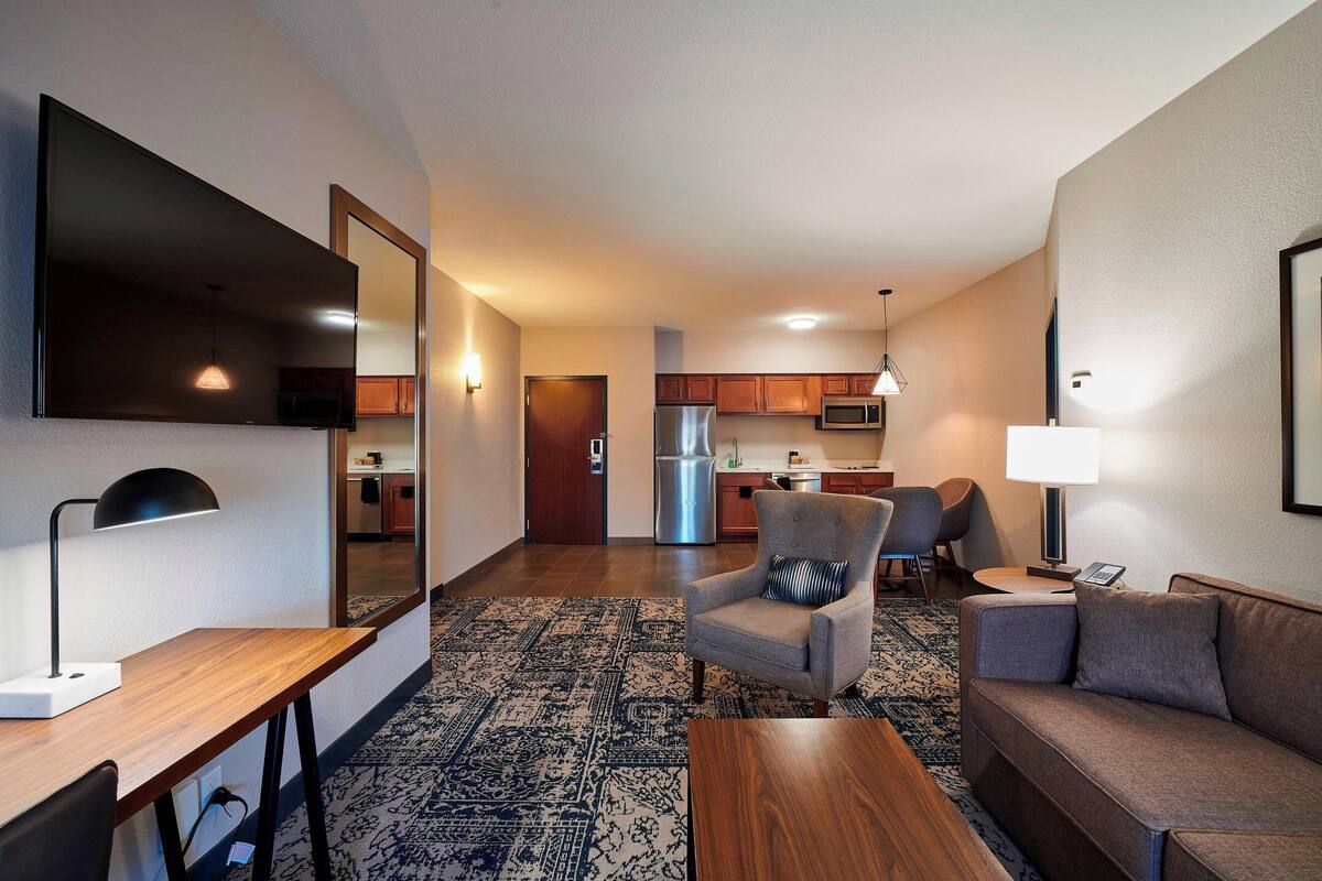 A modern hotel room with a flat-screen TV, a desk with a lamp, a sofa, a patterned rug, and a kitchenette in the background.