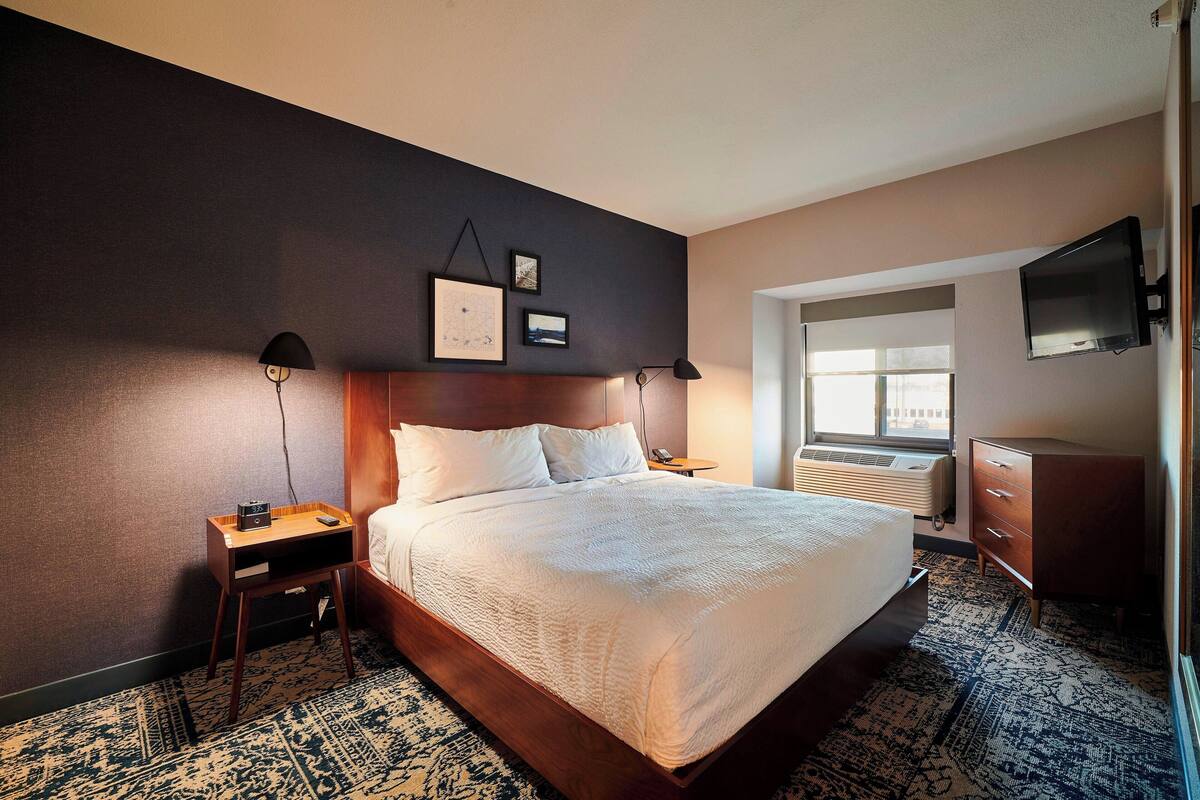 A hotel room with a large bed, a bedside table with a lamp, a dresser, and a TV mounted on the wall.
