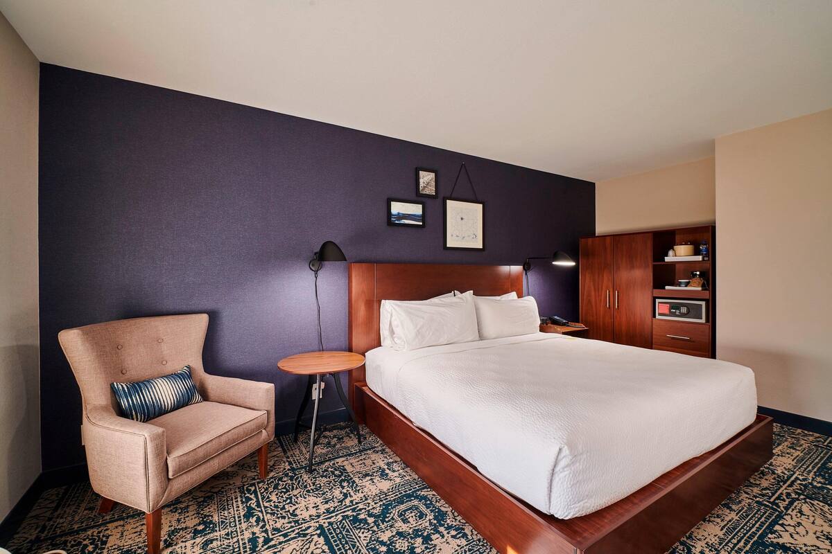 A hotel room with a large bed, a chair, a small table, and a wardrobe.