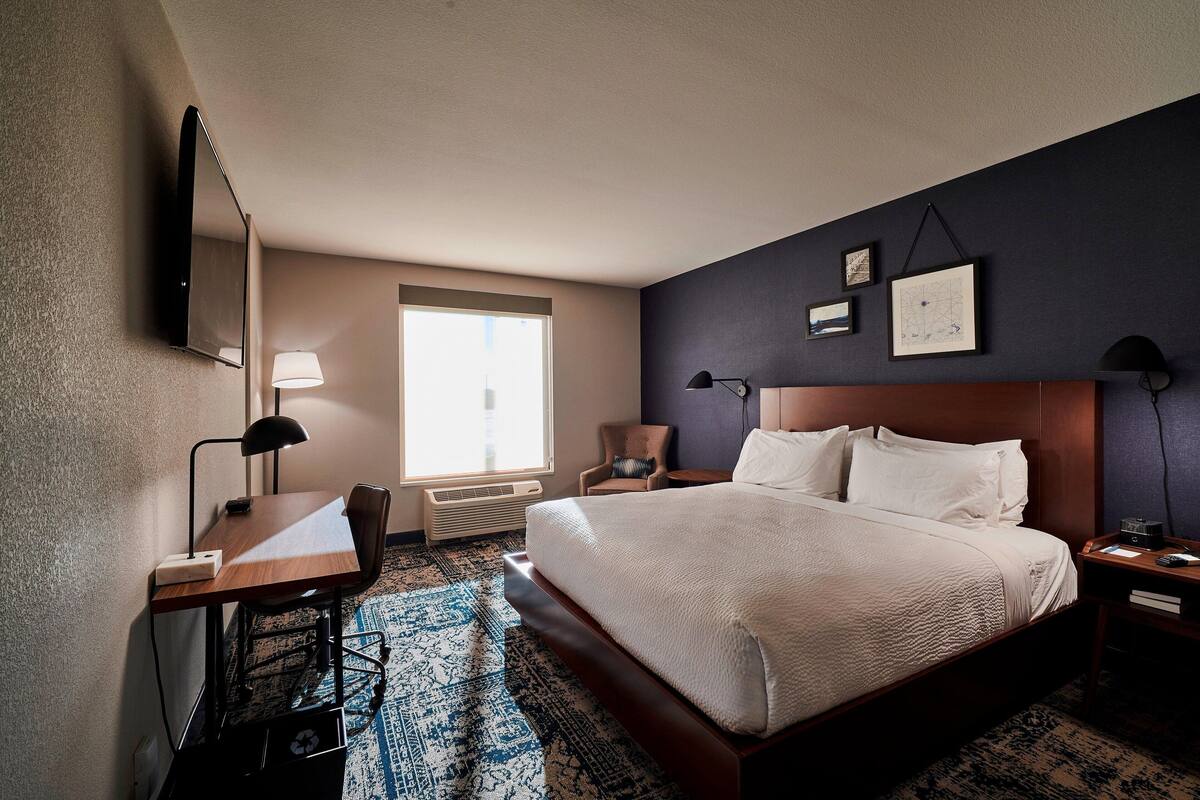 A hotel room with a large bed, a desk with a lamp, a chair, a nightstand, and a wall-mounted TV.