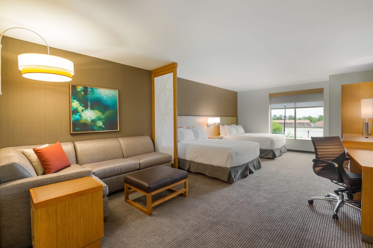 A hotel room with a sofa, two beds, a desk, and a large window.