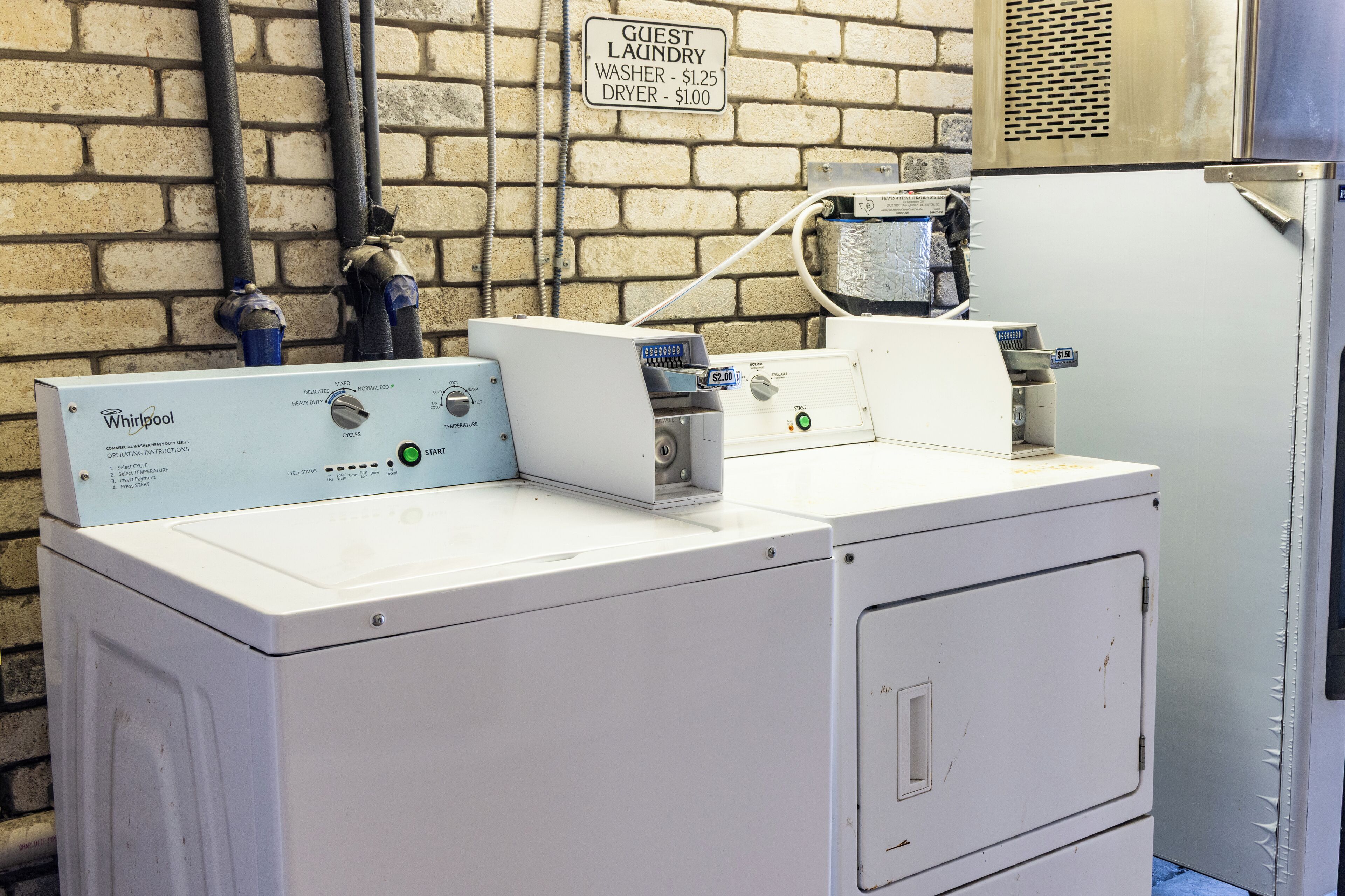 A Whirlpool washer and dryer in a utility room with a brick wall, a sign indicating guest laundry rates, and a water heater.