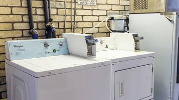 Une machine à laver et un sèche-linge Whirlpool se trouvent dans une buanderie, avec un mur en briques, un panneau indiquant les tarifs pour le service de blanchisserie et un chauffe-eau.