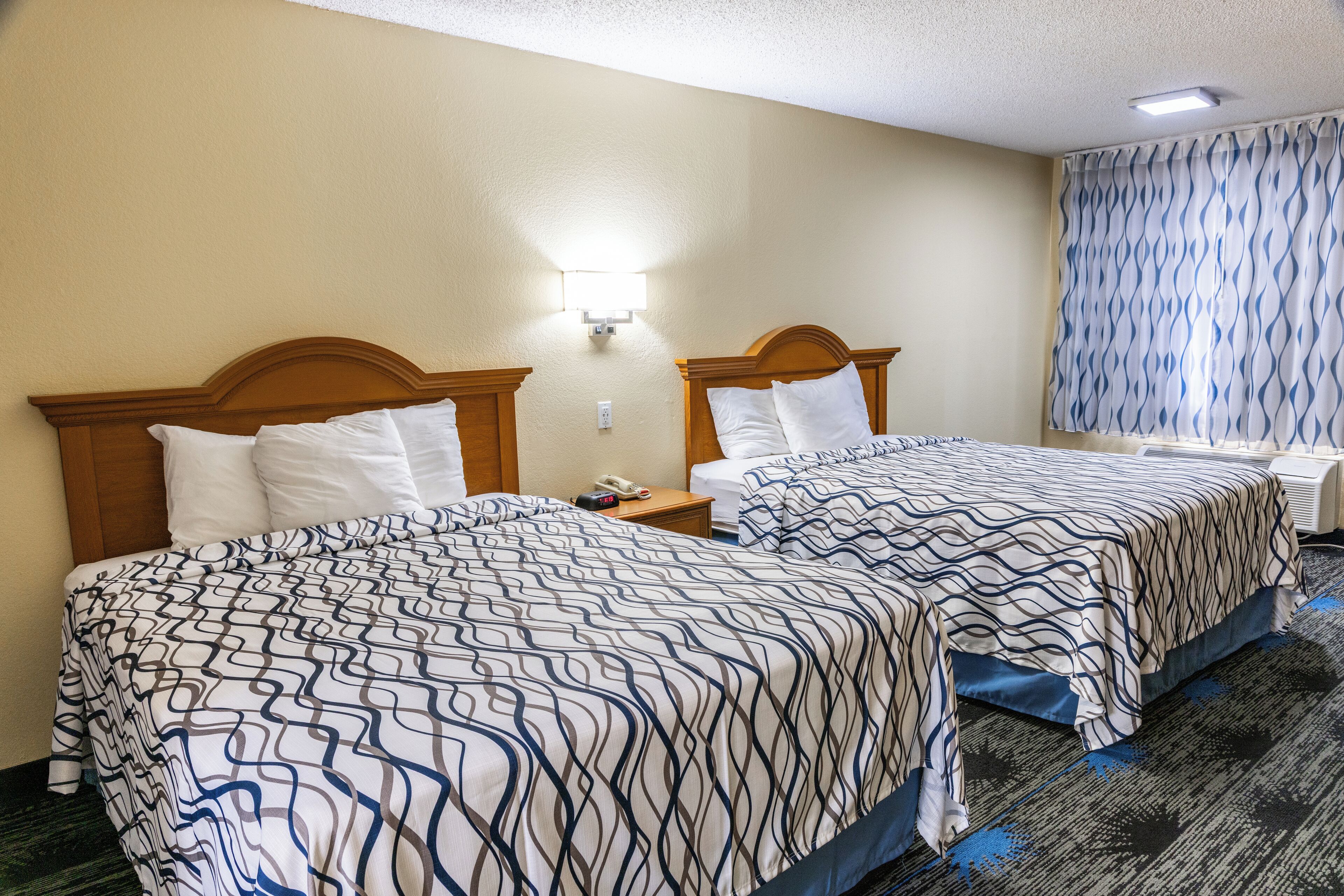 A hotel room with two beds, a nightstand, a lamp, and a window with patterned curtains.