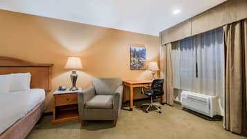 A hotel room with a bed, a desk, a chair, a lamp, a painting, and a window with curtains.