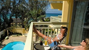 A person reclining on a balcony chair with a drink, overlooking a pool and ocean view. - Golden Sands Hotel (Oistins)