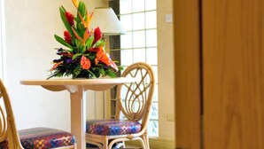 A dining area with a table and two wicker chairs, one with a colorful cushion. - Golden Sands Hotel (Oistins)