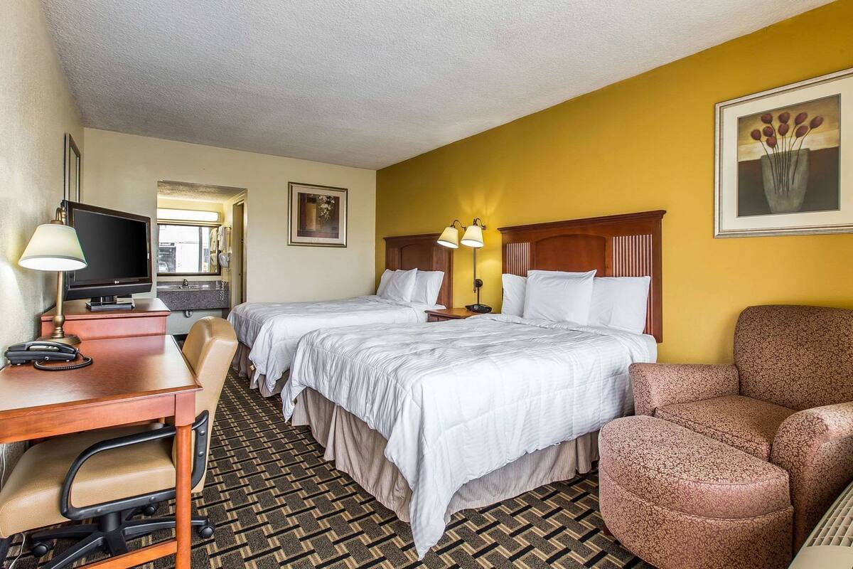 A hotel room with two beds, a desk with a lamp, a television, and a patterned carpet.