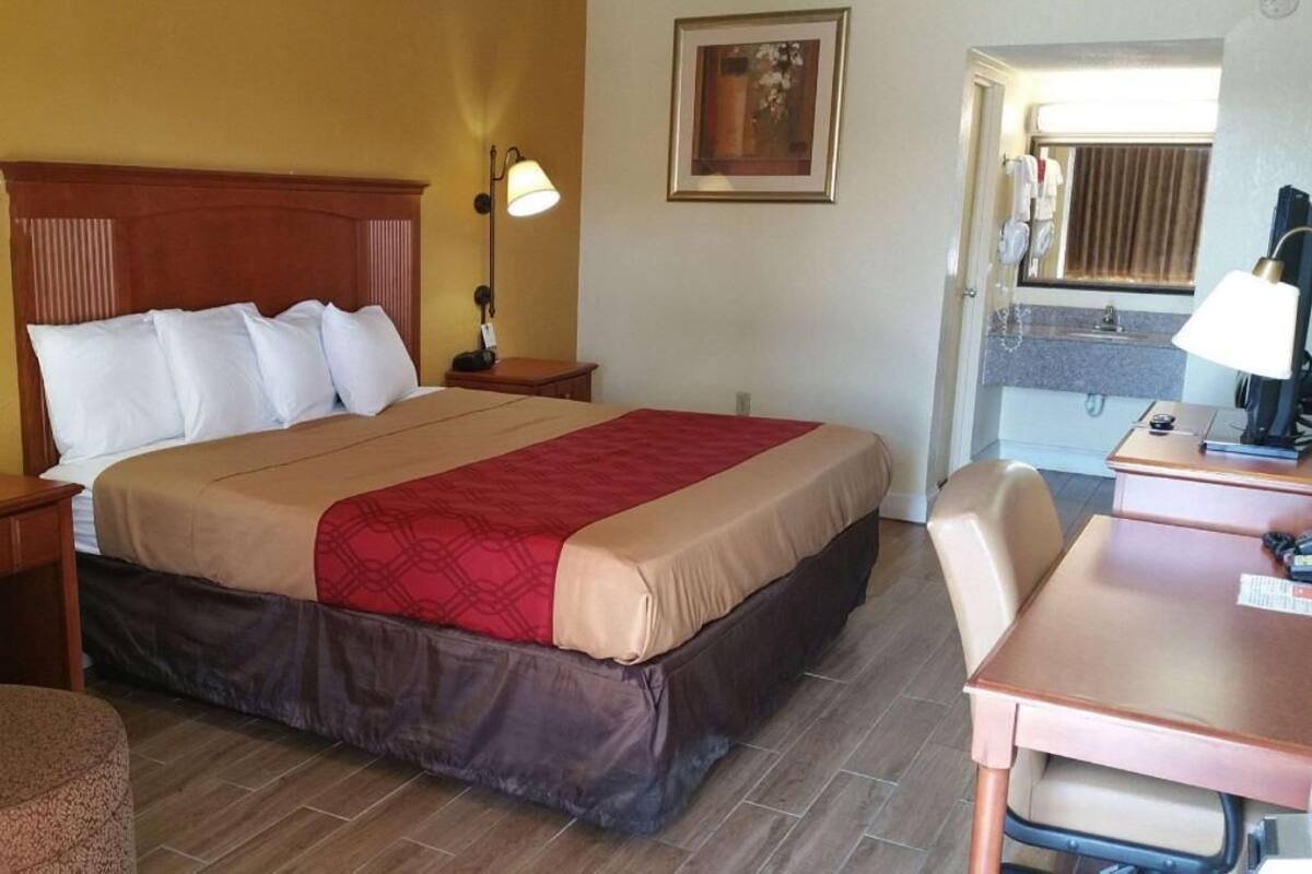 A hotel room with a bed, a desk, a chair, a nightstand, a lamp, a painting on the wall, and a bathroom visible through an open door.