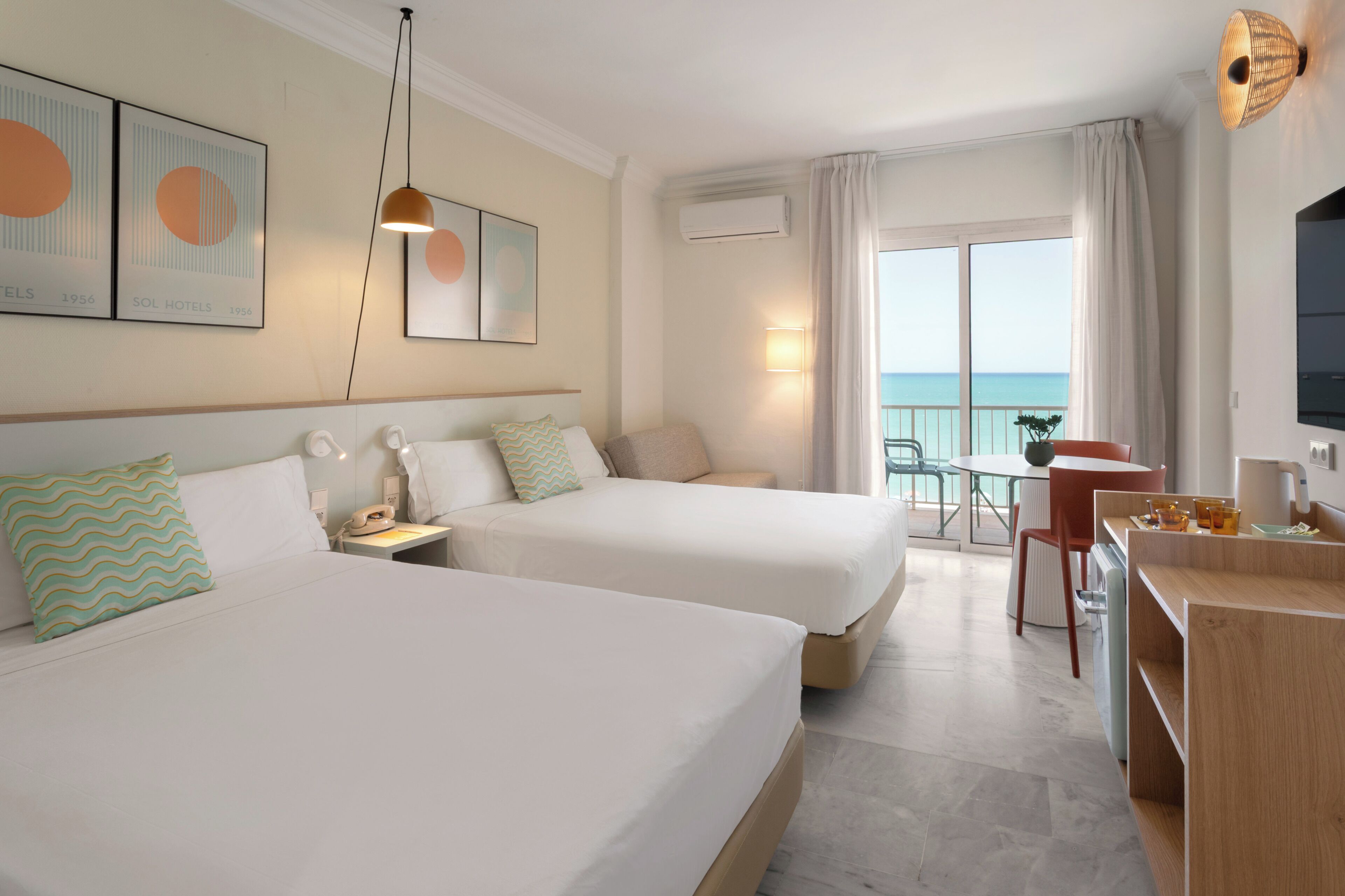 A hotel room with two beds, a desk with a chair, a TV, and a balcony with a view of the sea.