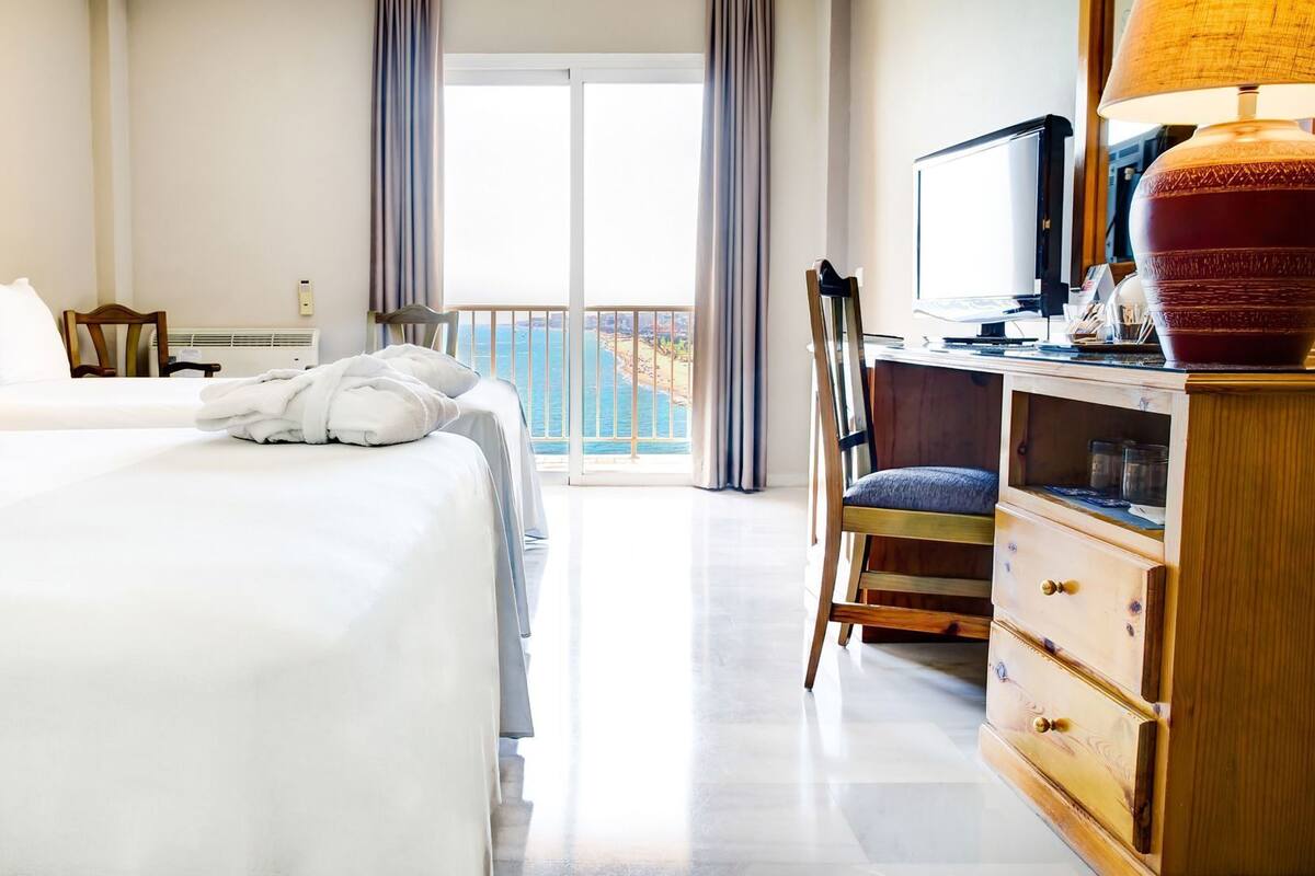 A hotel room with a bed, a desk, a chair, a TV, and a view of the sea through the window.