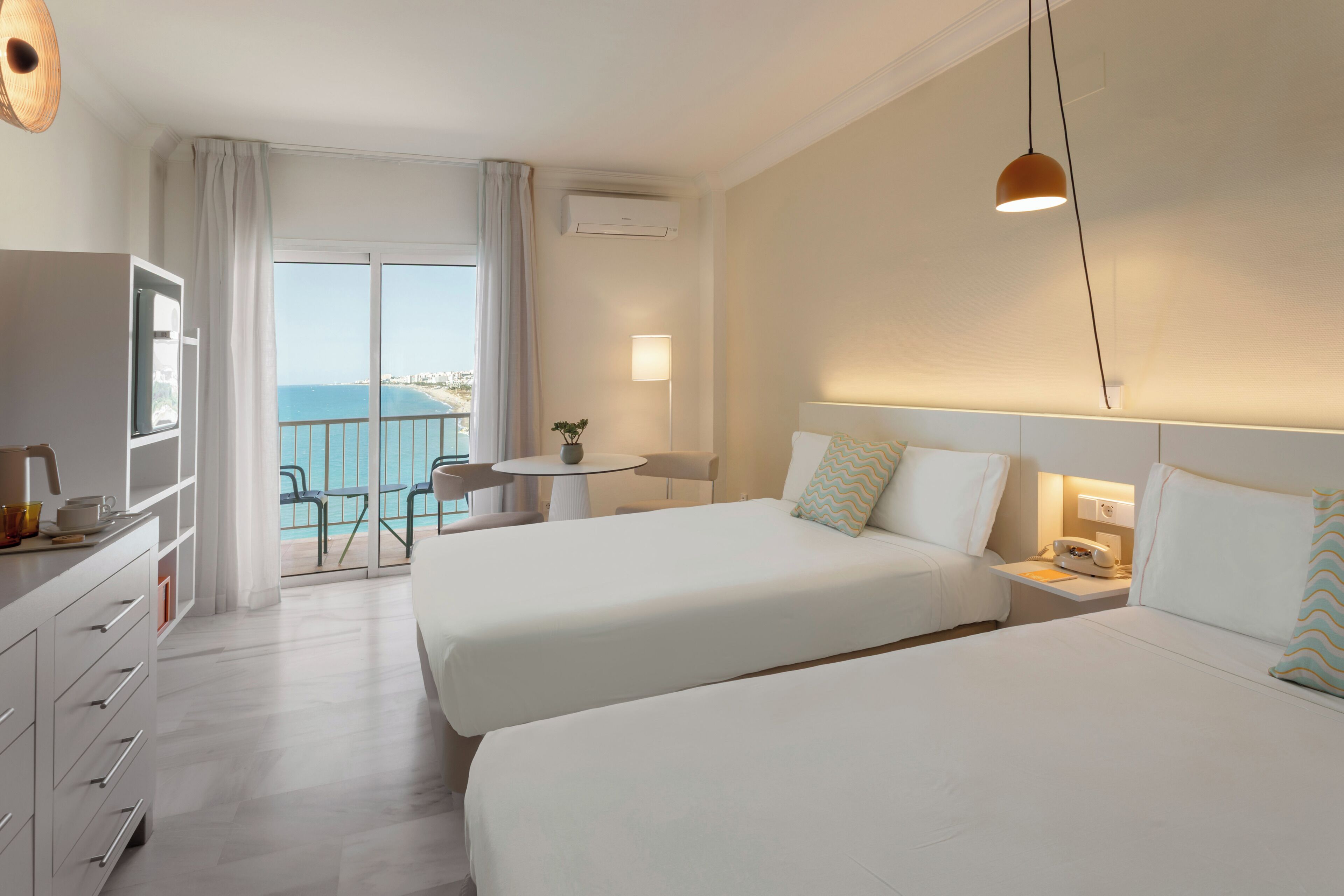 A hotel room with two beds, a desk, a chair, a small table with a plant, and a view of the sea.