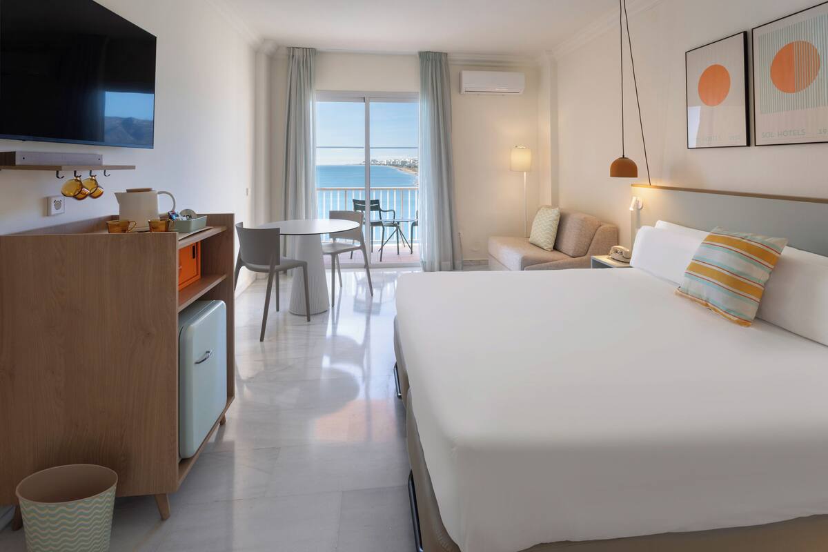 A hotel room with a bed, a desk with a computer, a chair, a small table, a sofa, and a balcony with a view of the sea.