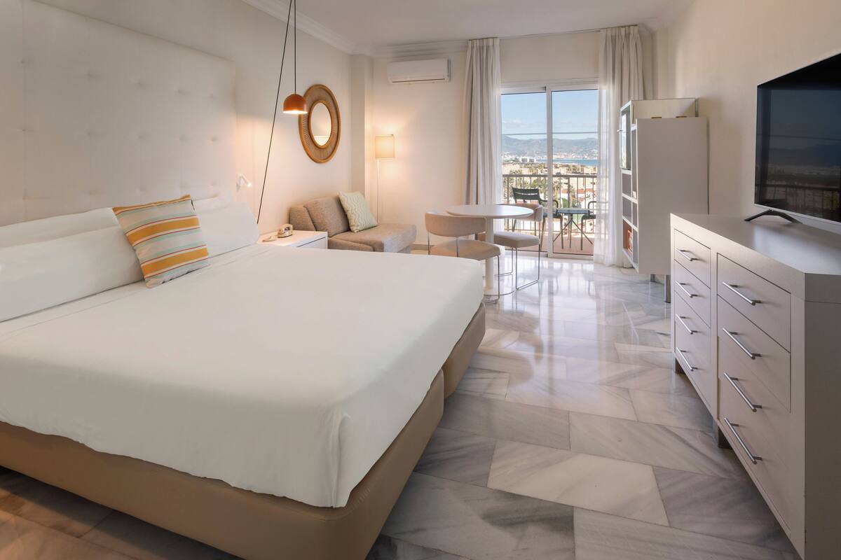 A modern hotel room with a large bed, a small sofa, a dining area with a table and chairs, and a TV on a dresser.
