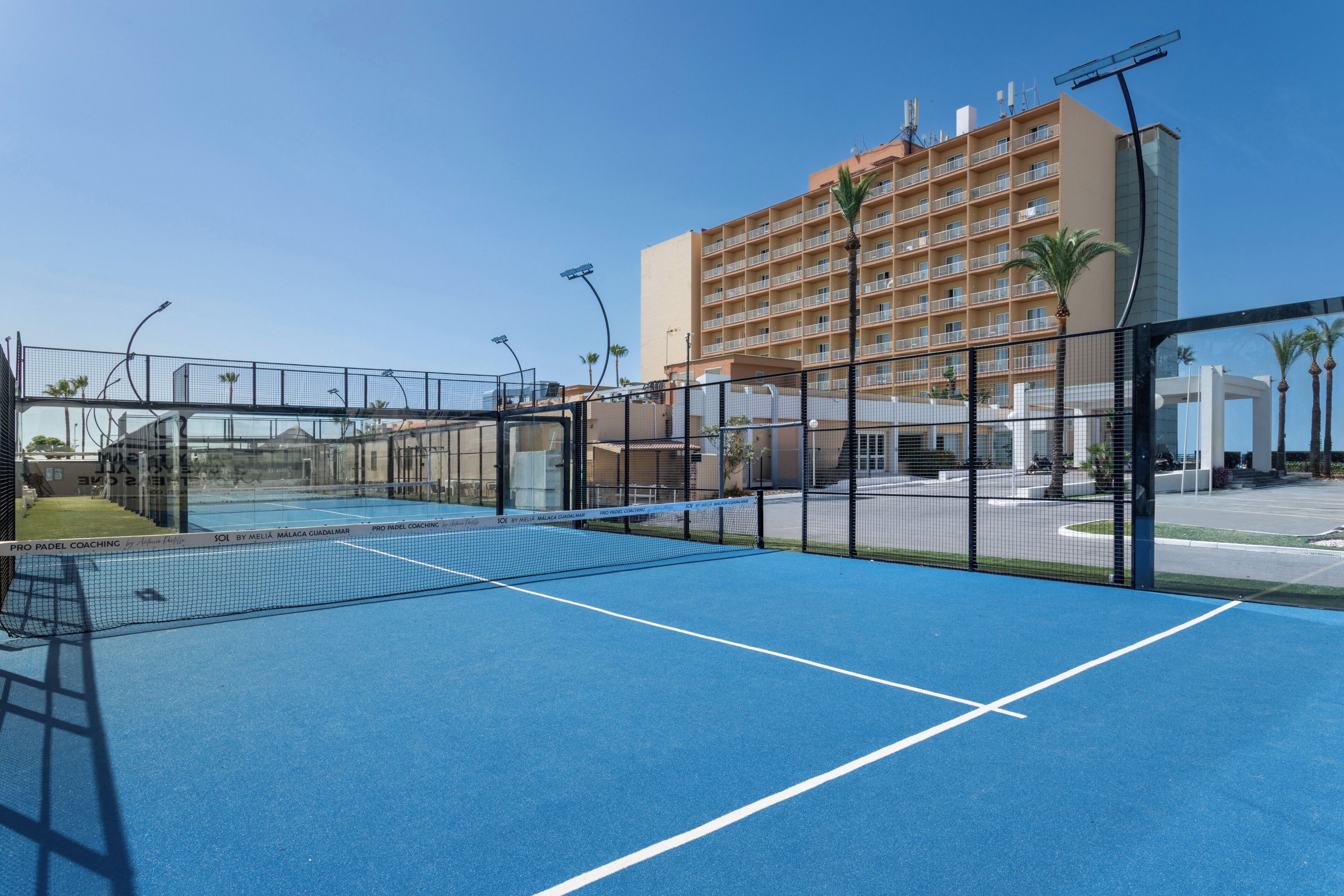 An outdoor tennis court with a net, surrounded by a fence, and a hotel building in the background.