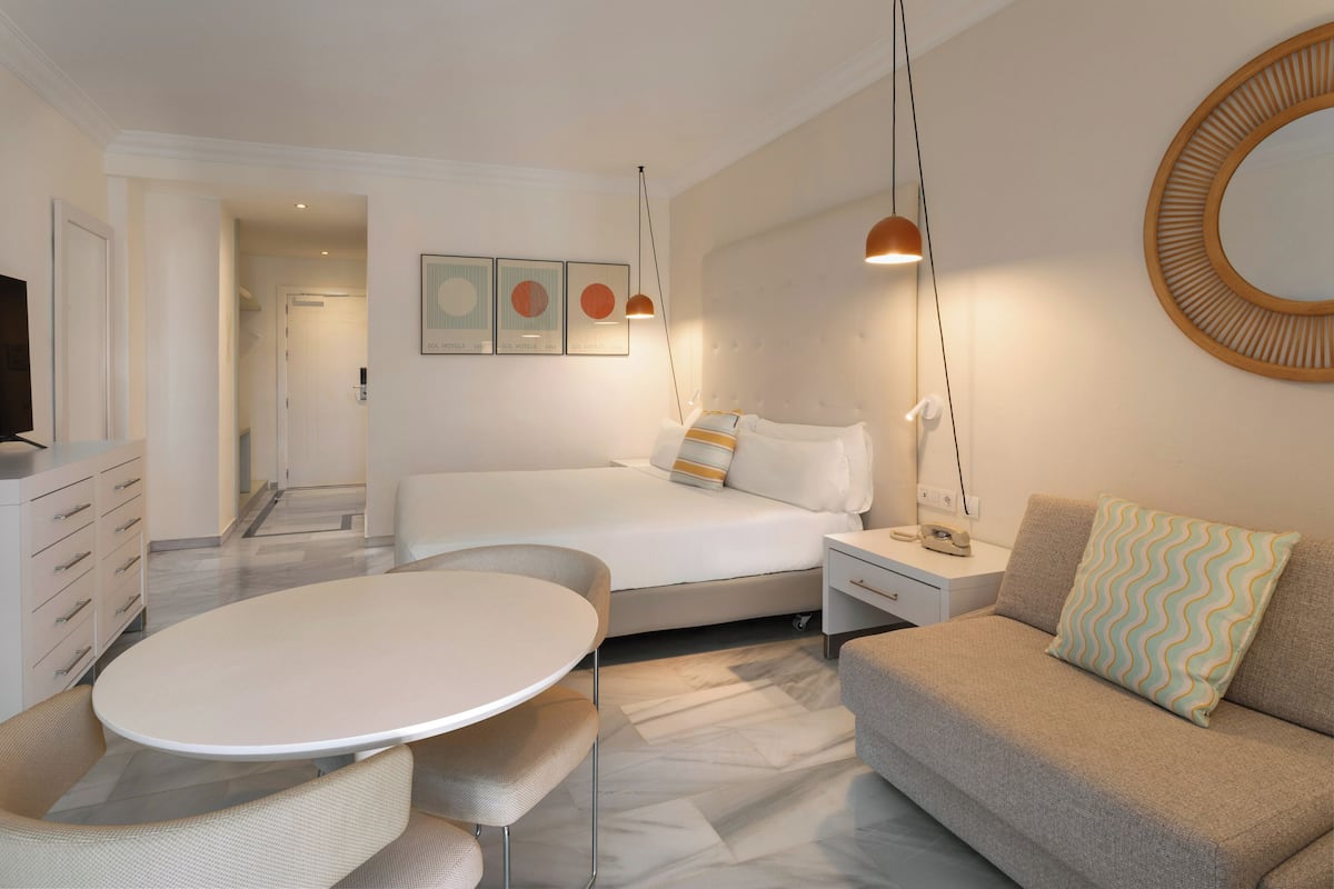 A modern hotel room with a bed, a sofa, a dining table, and a TV.