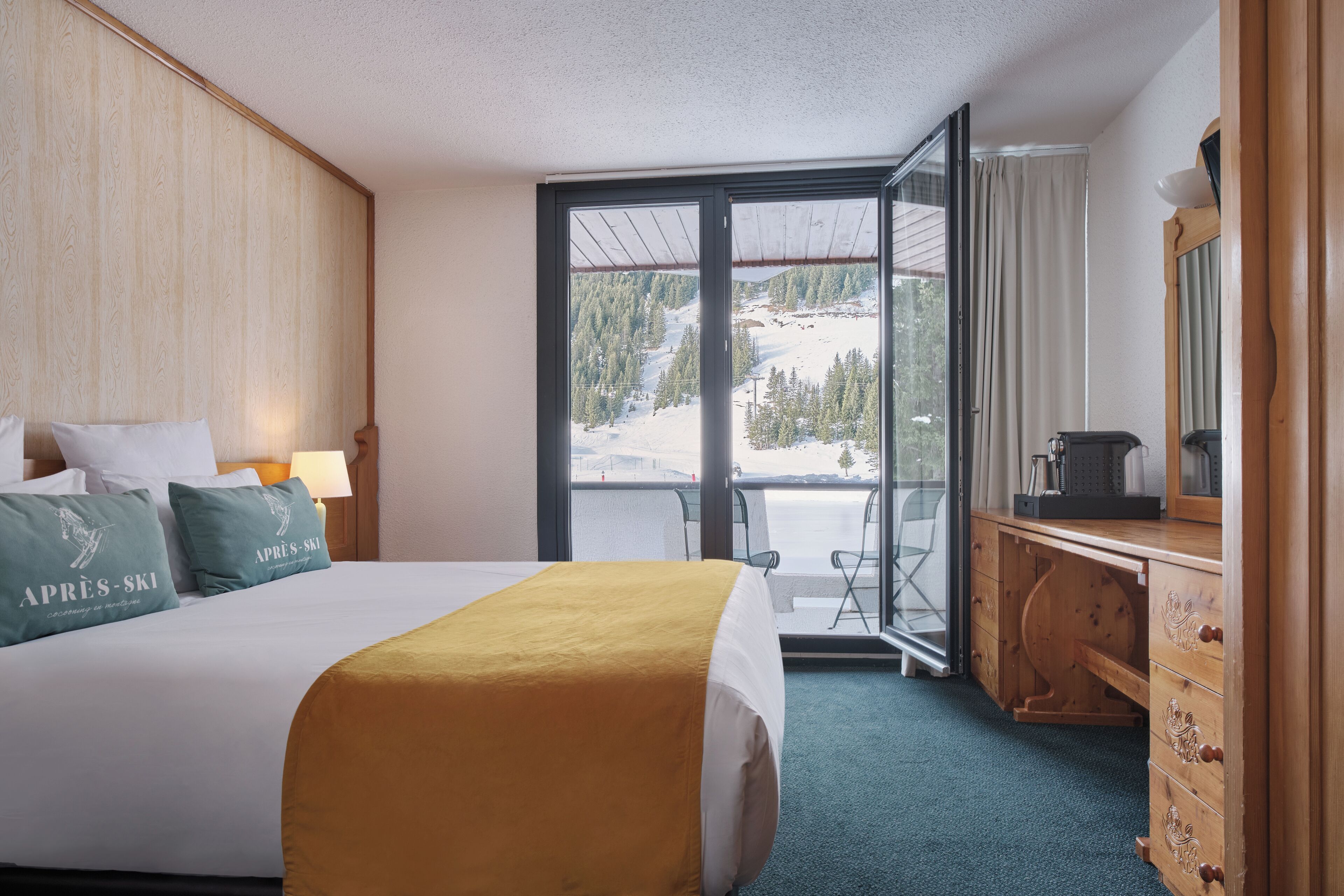 Une chambre d’hôtel avec un lit, une commode en bois, un bureau avec un ordinateur et une grande fenêtre donnant sur une montagne enneigée.