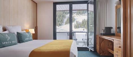Habitación de hotel con cama, cómoda de madera, escritorio con computadora y un amplio ventanal con vistas a una montaña nevada.
