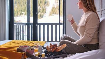 Una persona sentada en una cama con una bandeja de desayuno, que incluye cruasanes, un bol de bayas y un vaso de zumo de naranja, con vistas a un paisaje nevado a través de una ventana.