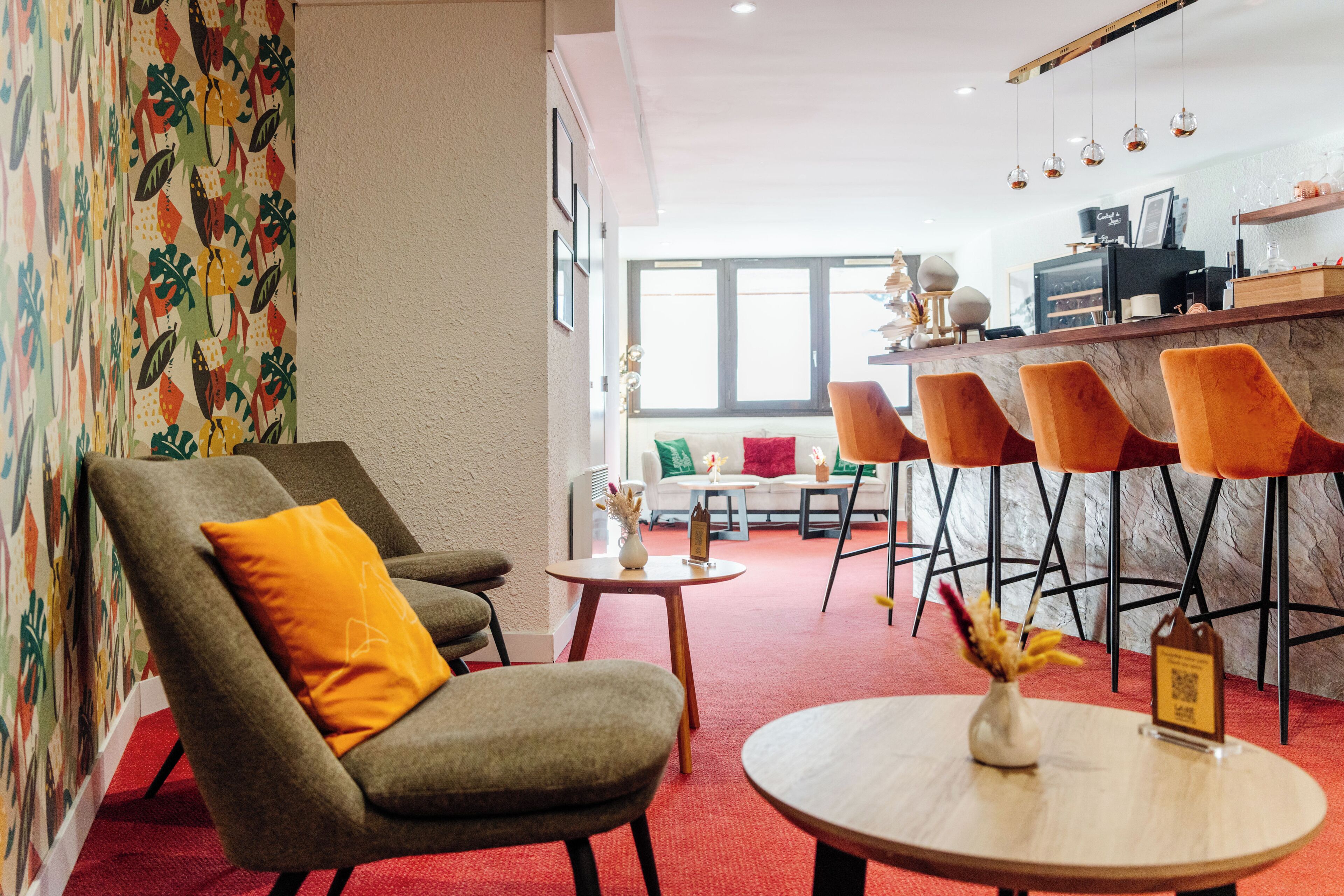 A modern hotel lobby with a red carpet, a bar area with orange chairs, and a seating area with a grey sofa and an orange pillow.