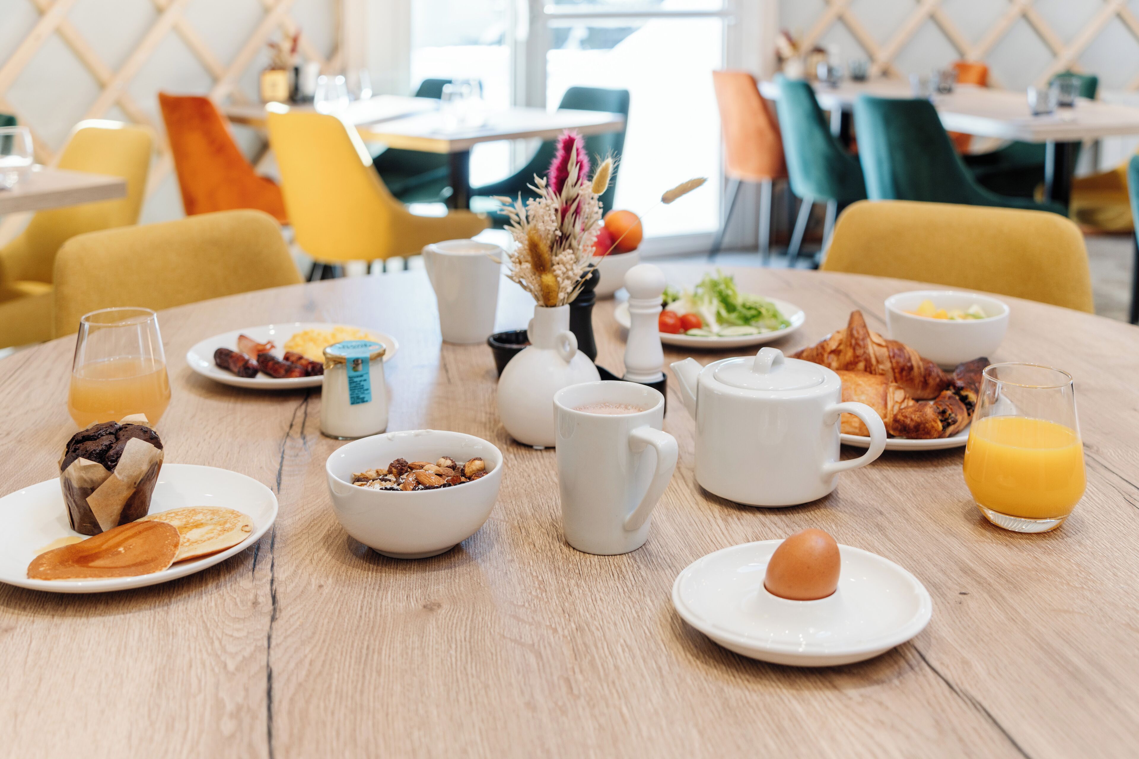 Une table en bois dressée pour le petit-déjeuner, avec différents plats, notamment des crêpes, des œufs et des toasts.