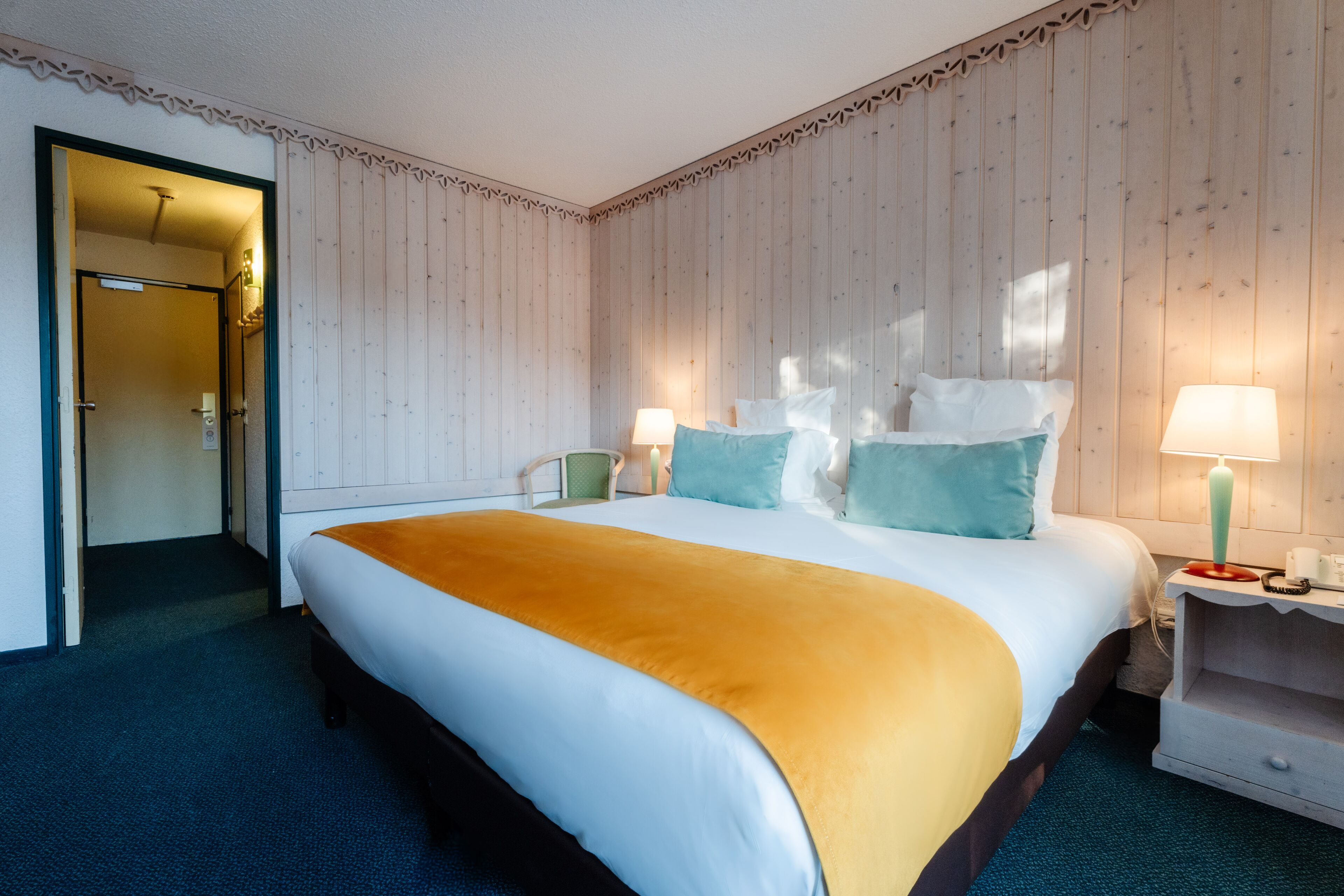 Habitación de hotel con una cama grande, dos mesitas de noche, una silla y una puerta que da a otra habitación.