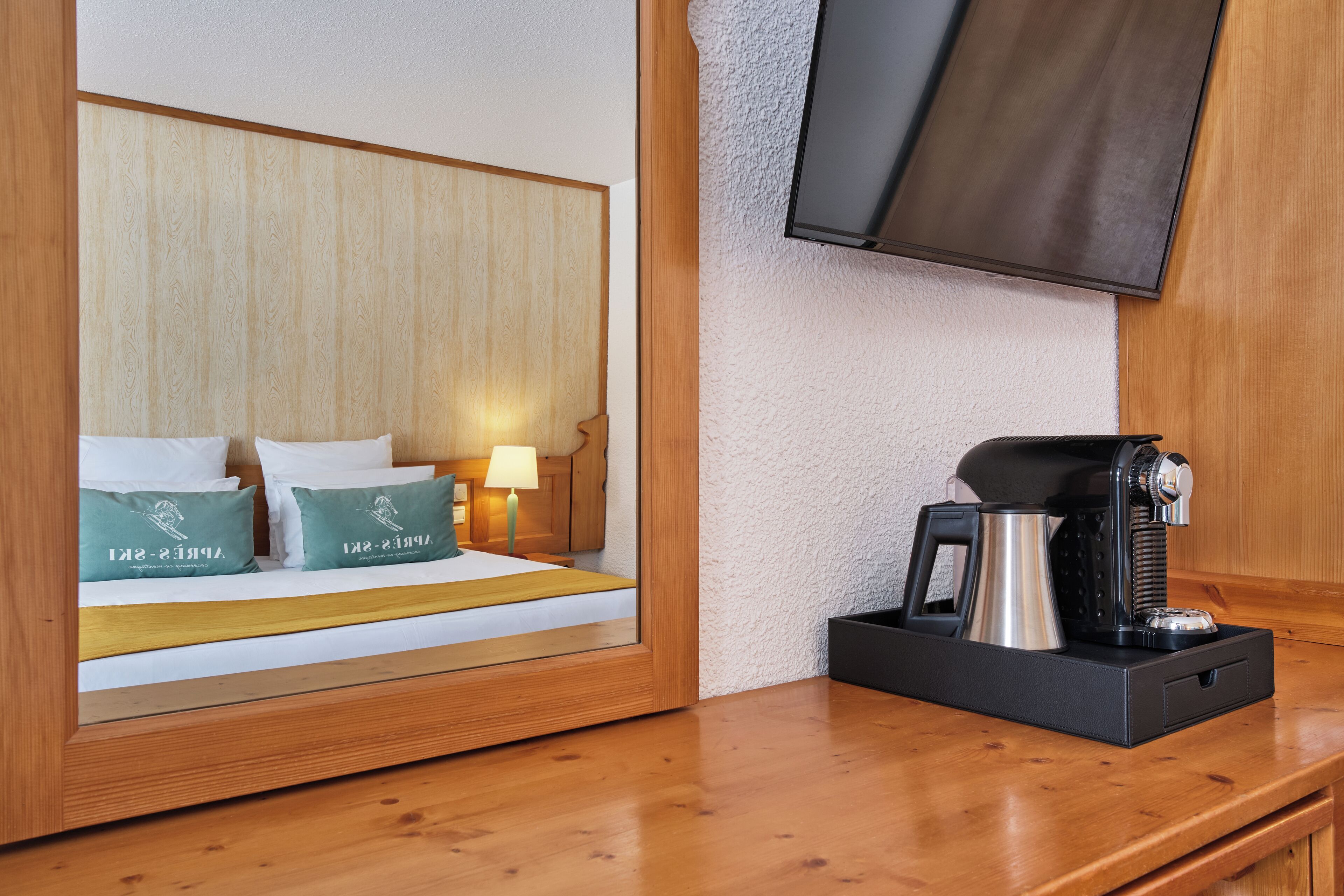Habitación de hotel con cabecera de madera, cama con almohadas verdes, lámpara de noche y cafetera en la mesita.