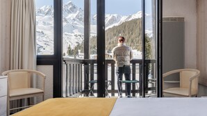 A room with a large window offering a mountain view, a bed, two chairs, and a wooden railing. - Lake Hôtel Courchevel 1850 (Courchevel)