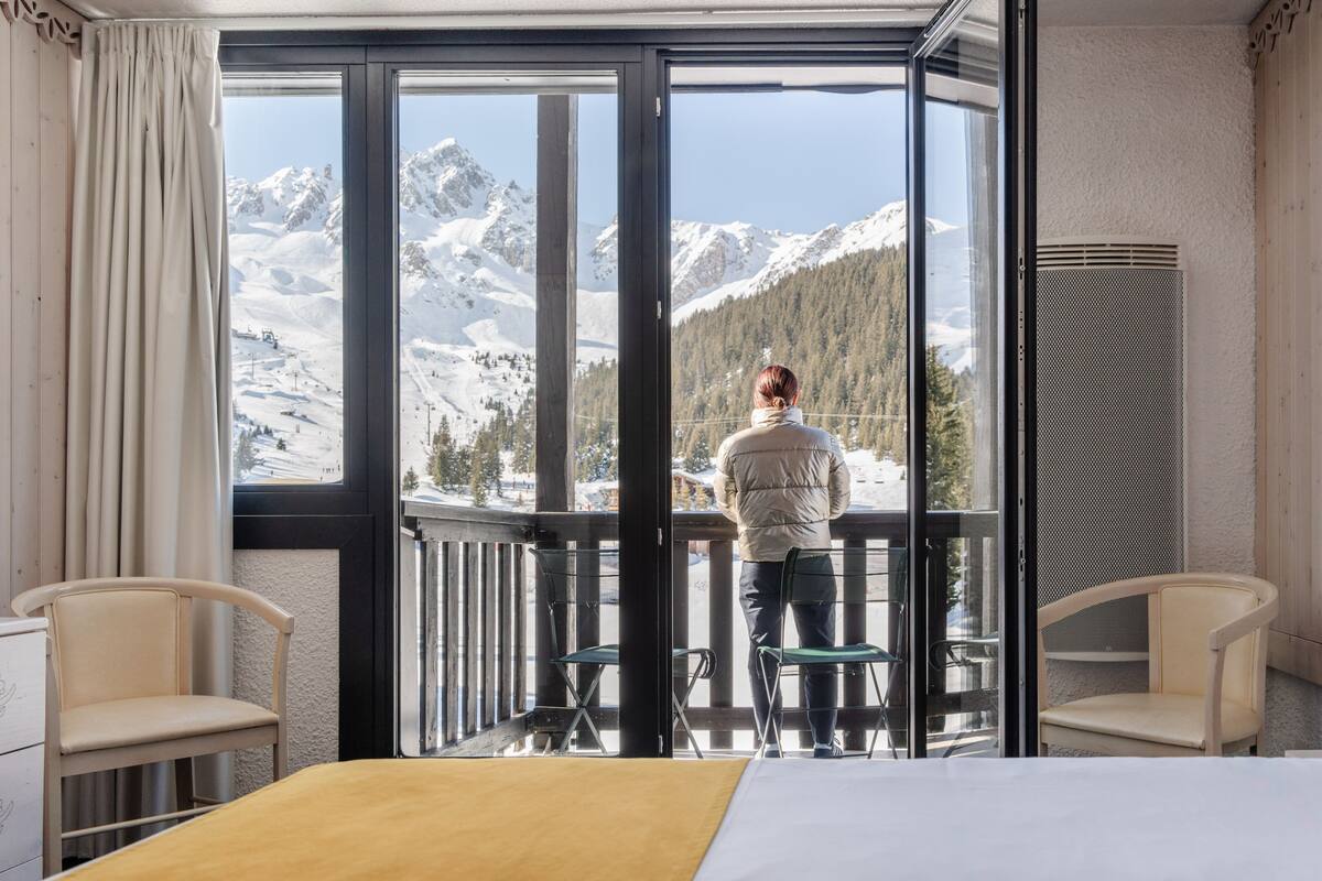 A room with a large window offering a mountain view, a bed, two chairs, and a wooden railing.