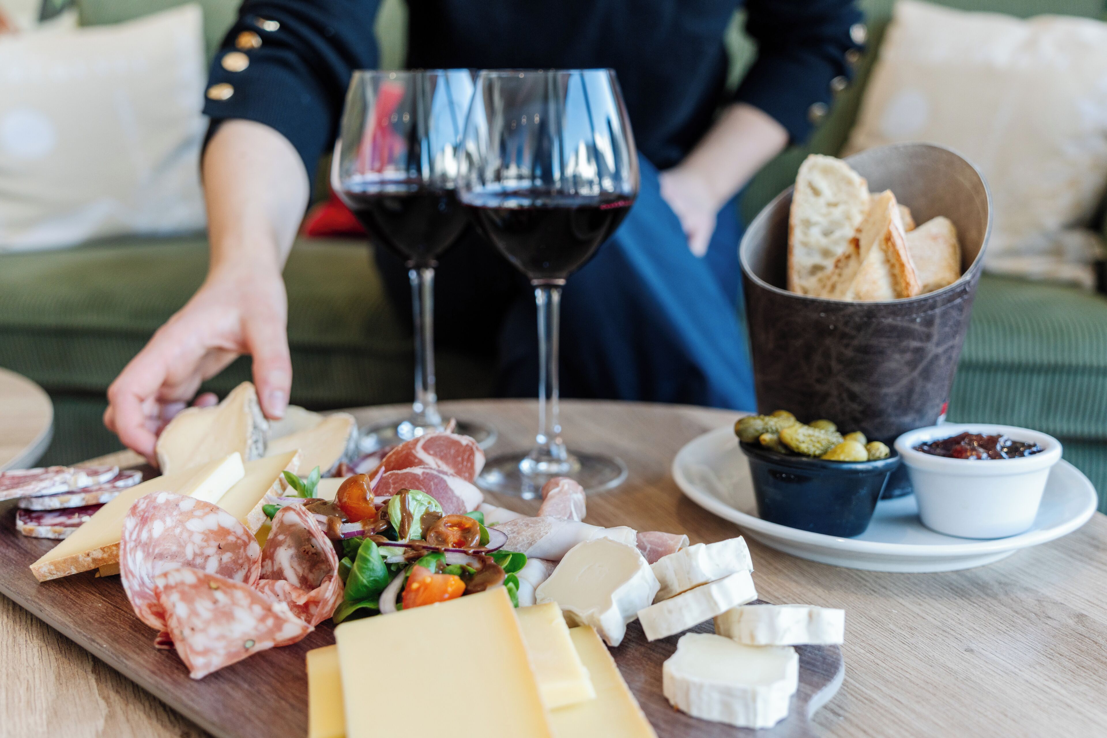 Un tagliere con formaggi, salumi e pane assortiti, accompagnato da bicchieri di vino e una tazza di caffè.