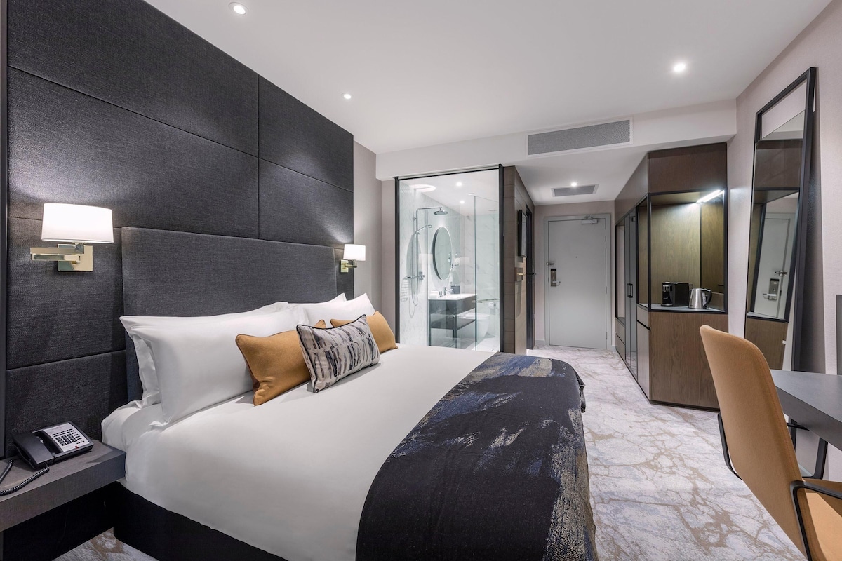 A modern hotel room with a large bed, bedside lamps, a desk, and a bathroom visible through a glass door.