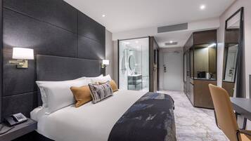 A modern hotel room with a large bed, bedside lamps, a desk, and a bathroom visible through a glass door.
