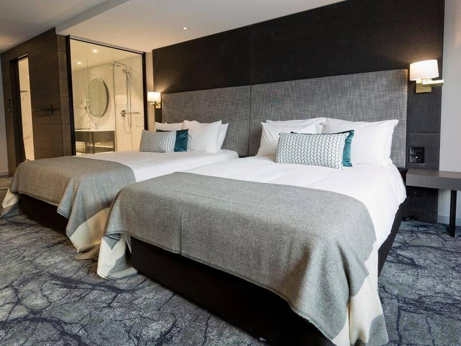 A hotel room with two beds, a glass shower enclosure, and a bedside table with a lamp.