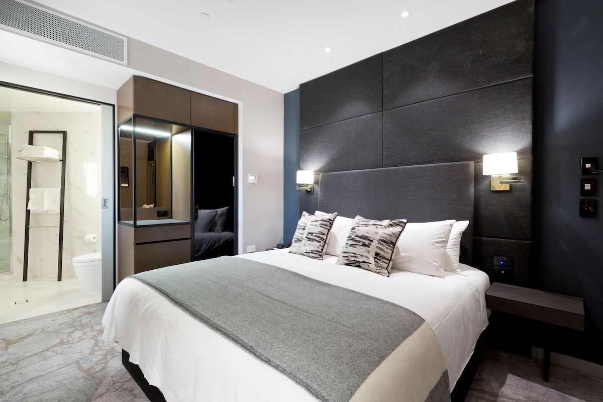 Une chambre d’hôtel moderne dotée d’un grand lit, d’une table de chevet, d’une commode, d’un miroir et d’une salle de bain avec une douche à parois de verre.