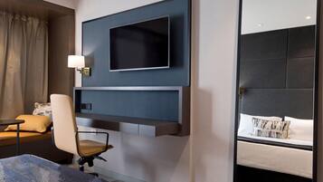 Een hotelkamer met een wandgemonteerde flatscreen-televisie, een bureau met stoel, een bed met kussens en een wandlamp.