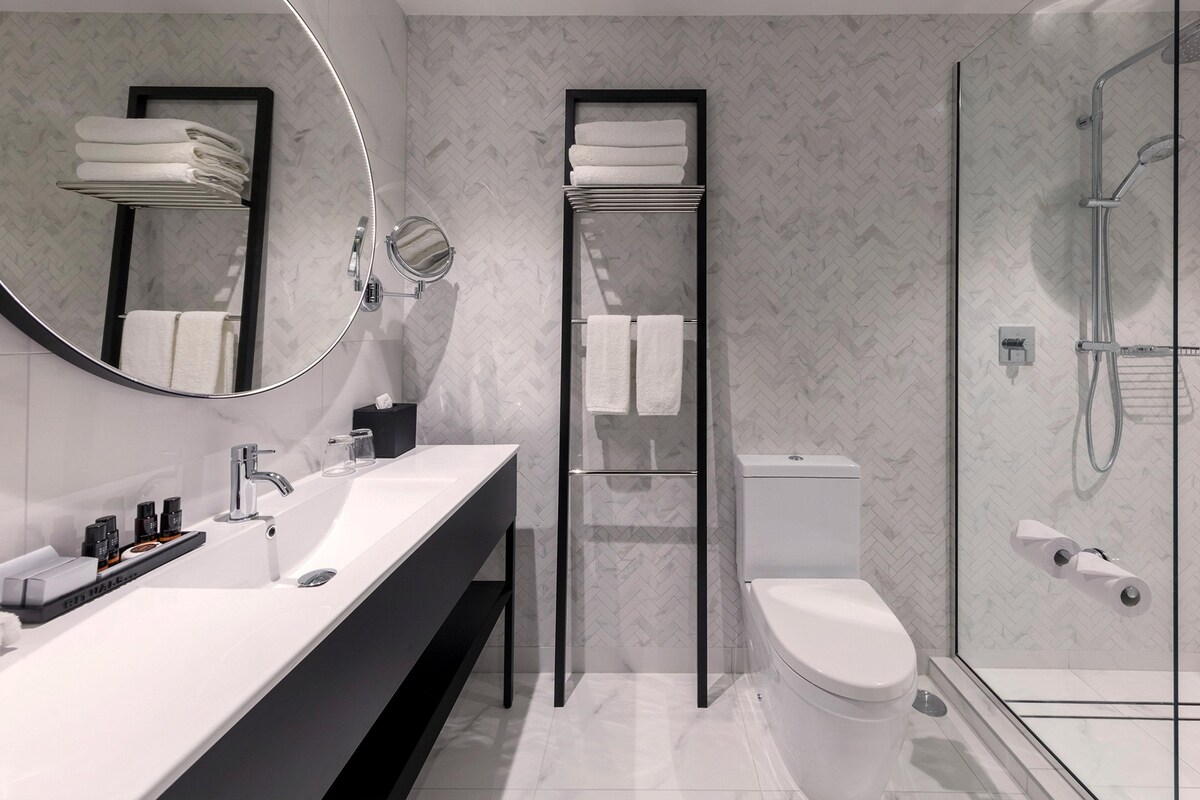 Ein modernes Badezimmer mit weißem Waschbecken, Toilette, Duschkabine aus Glas und einem Spiegel mit Handtuchhalter.