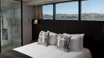Habitación de hotel moderna con una cama grande, dos almohadas decorativas, cabina de ducha de vidrio y vistas a un paisaje urbano a través de amplios ventanales.