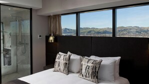 A modern hotel room with a large bed, two decorative pillows, a glass shower enclosure, and a view of a cityscape through large windows. - Crowne Plaza Christchurch (Christchurch)