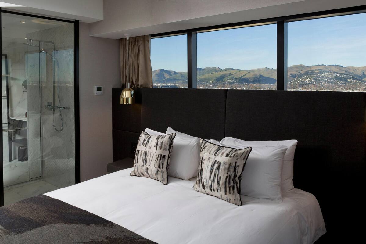 Ein modernes Hotelzimmer mit einem großen Bett, zwei Zierkissen, einer Glasdusche und Blick auf eine Stadtlandschaft durch große Fenster.