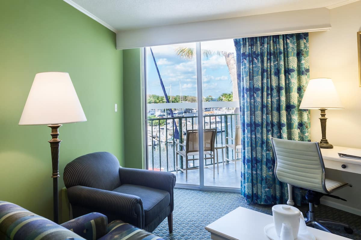 Habitación de hotel con sofá, escritorio, silla, lámpara y vistas a la marina a través de amplias ventanas.