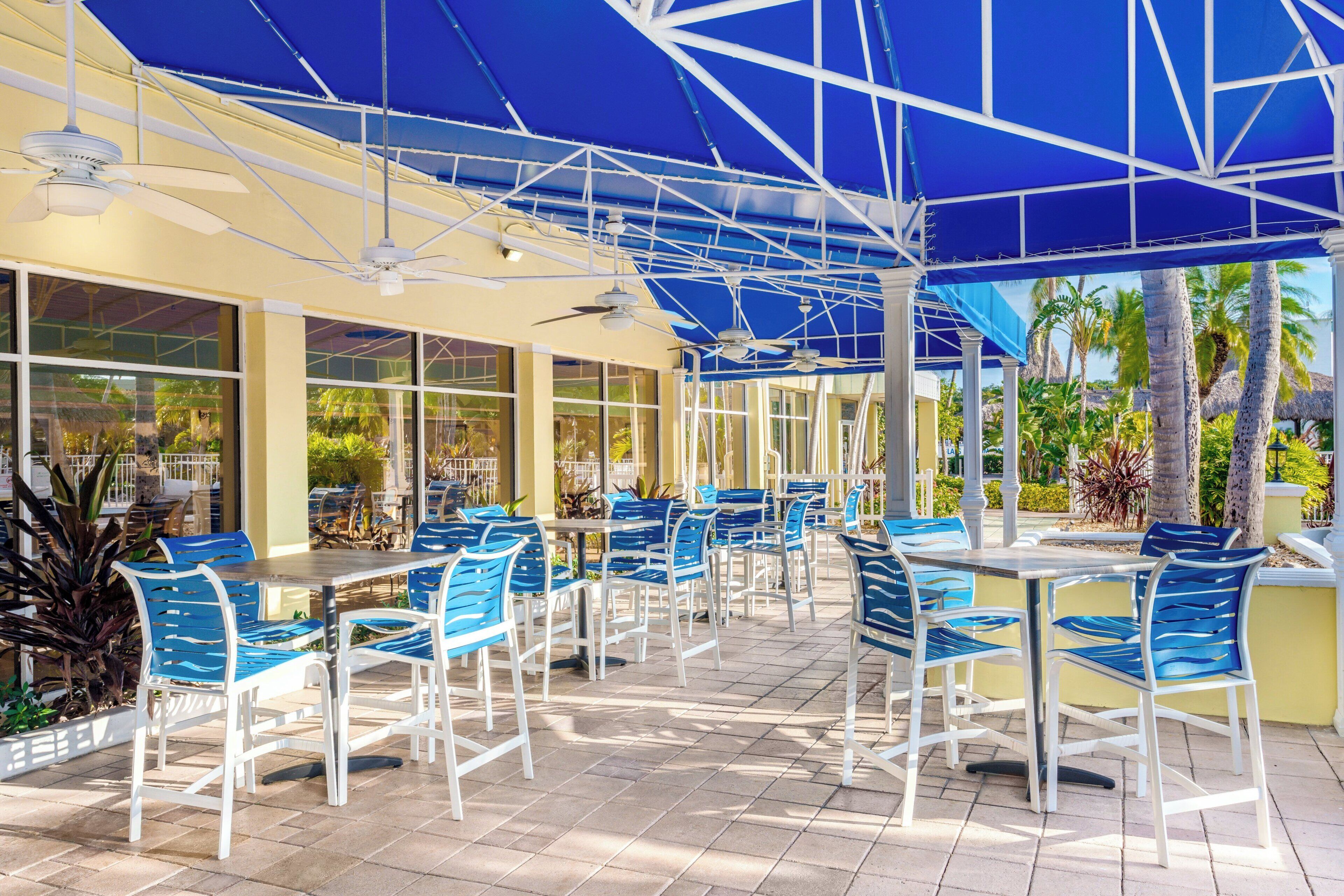 Un espace extérieur aménagé en terrasse, avec des tables et des chaises, de grandes fenêtres et une structure de tonnelle bleue.