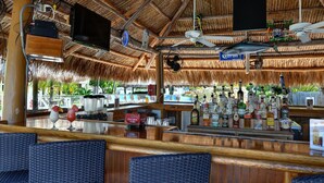 A bar area with a thatched roof, wooden furniture, and a variety of bottles.