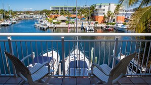 A balcony with two white chairs overlooking a marina with boats, a building with a green roof, and palm trees.