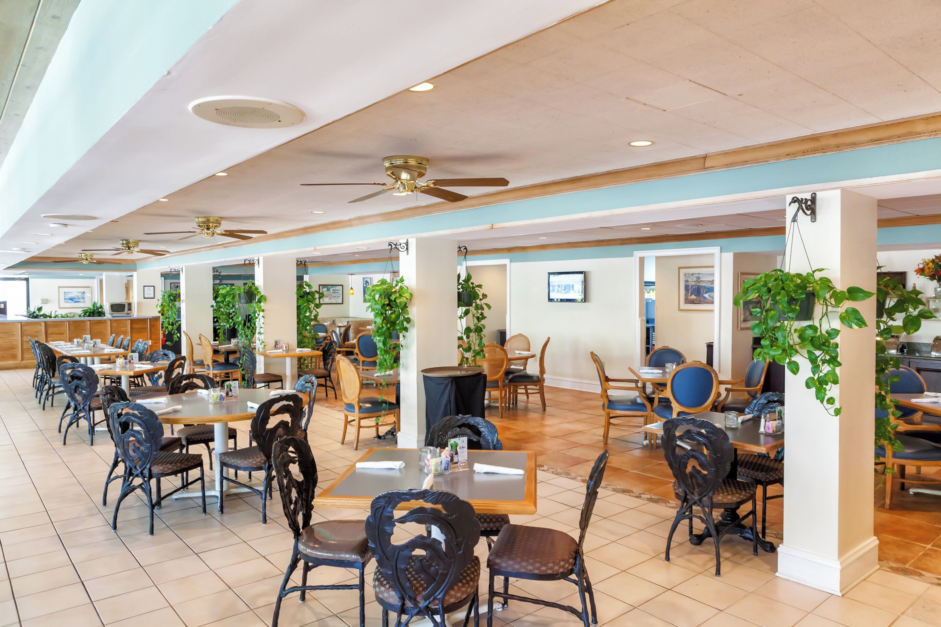 A spacious indoor dining area with multiple tables and chairs, ceiling fans, and potted plants.