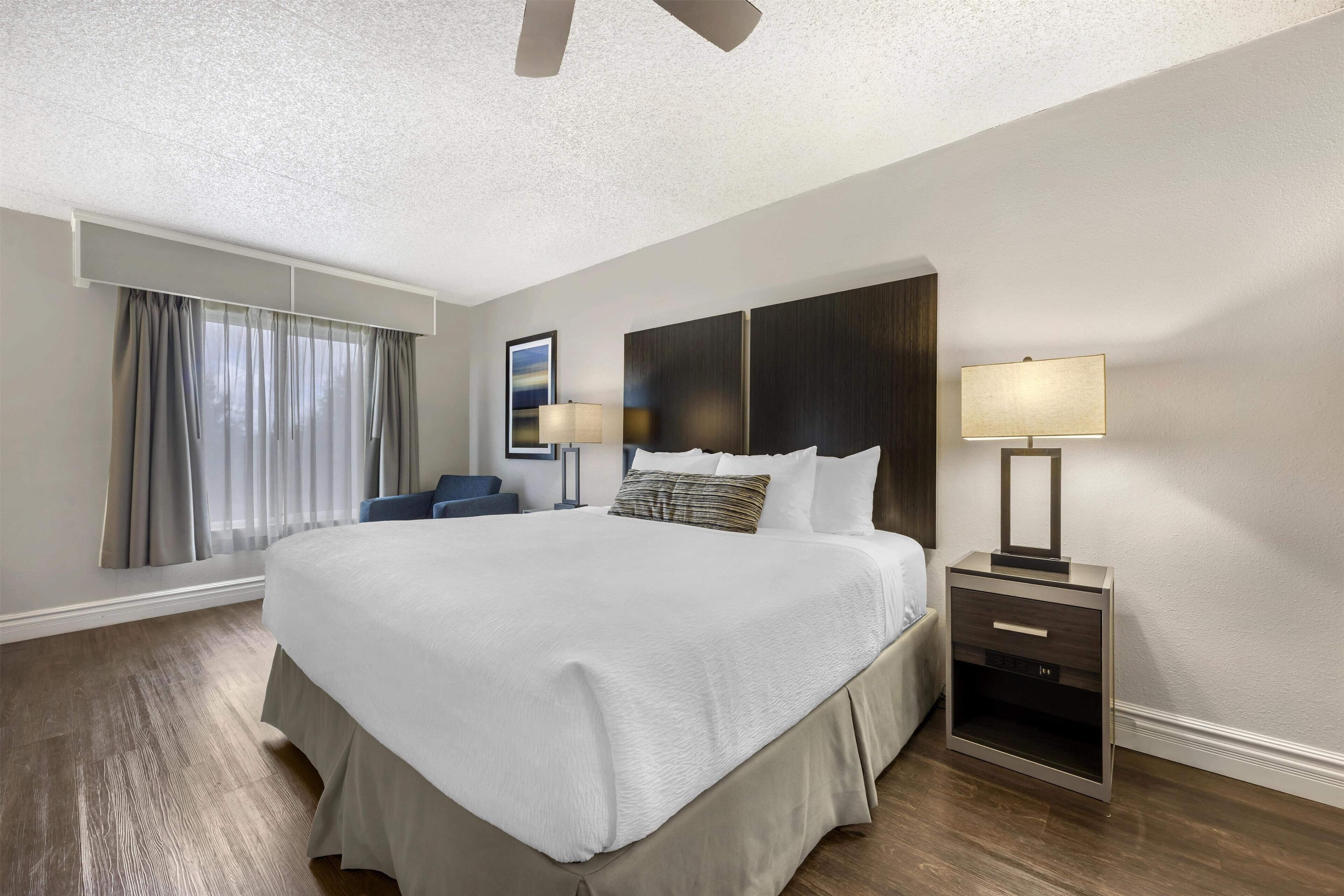 Habitación de hotel con una cama grande, dos mesitas de noche con lámparas, un ventilador de techo, una ventana con cortinas y un cuadro en la pared.