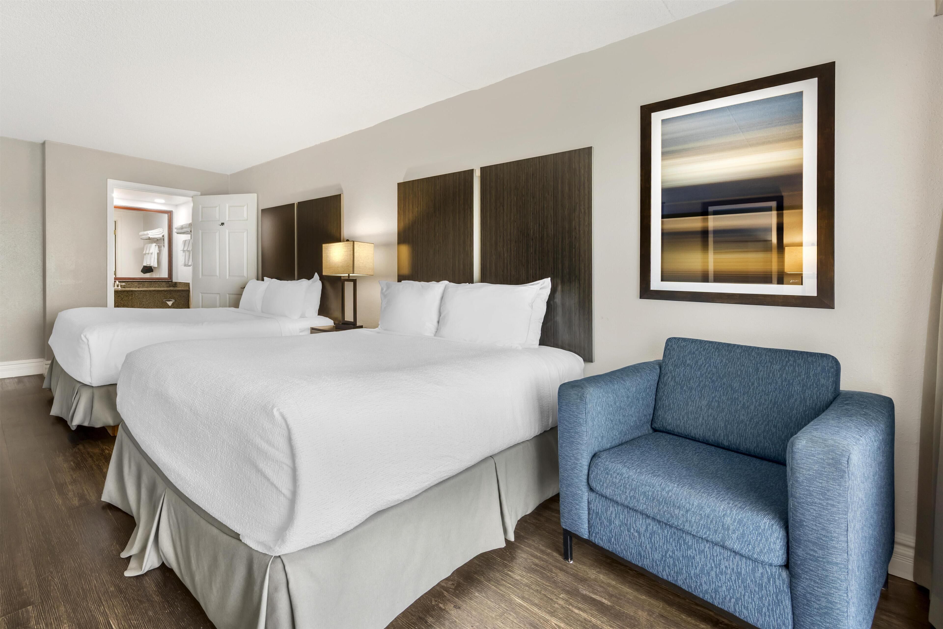 A hotel room with two beds, a blue armchair, a nightstand with a lamp, and a framed picture on the wall.