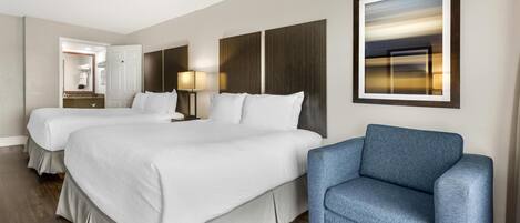 Habitación de hotel con dos camas, un sillón azul, una mesita de noche con lámpara y un cuadro en la pared.