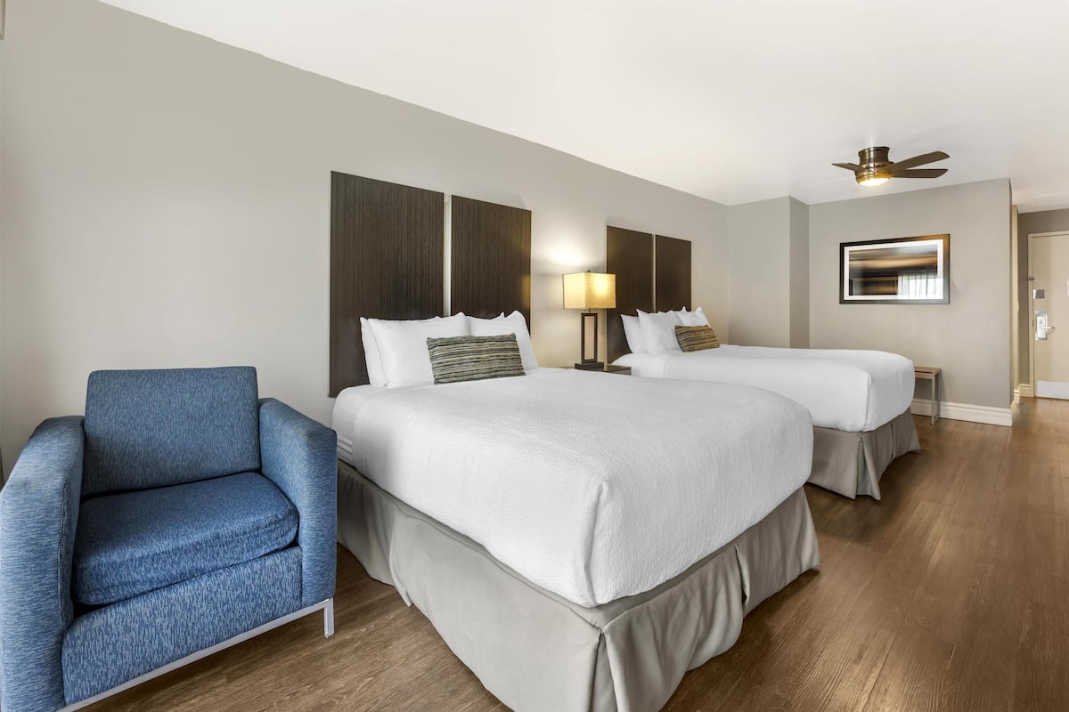 Una habitación de hotel con dos camas, un sillón azul, un ventilador de techo y una lámpara de pared.
